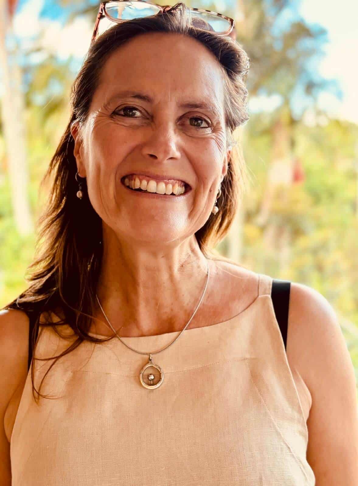 Smiling woman outdoors with sunglasses on her head and a beige top.