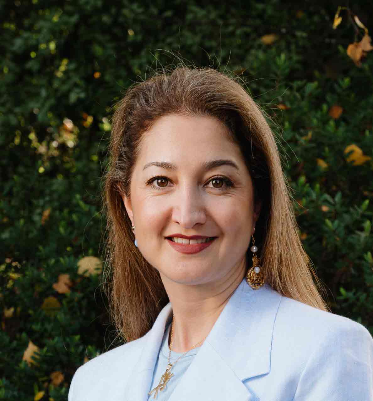 Portrait of a woman outdoors with greenery in the background, smiling, wearing a white blazer.