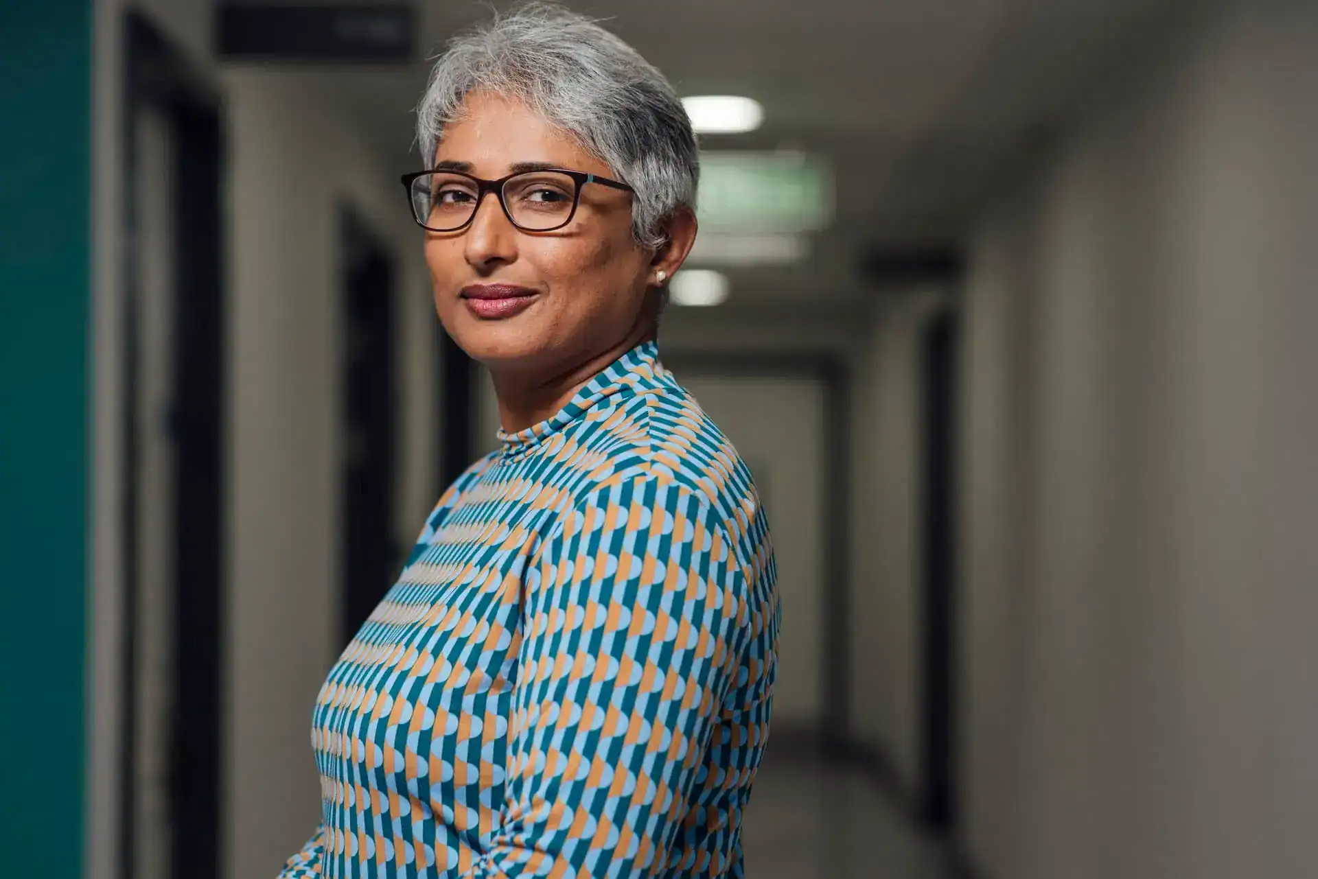 Professional woman with short grey hair and glasses in a corridor.